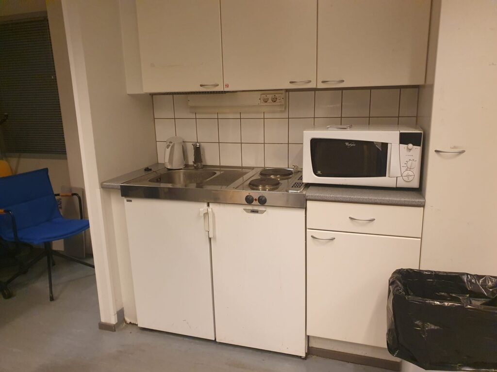 Espoolaisen toimistotilan vanha taukotilan keittiö ennen pintaremonttia: valkoiset kaapit, kaakeloitu välitila ja kaksilevyinen liesi